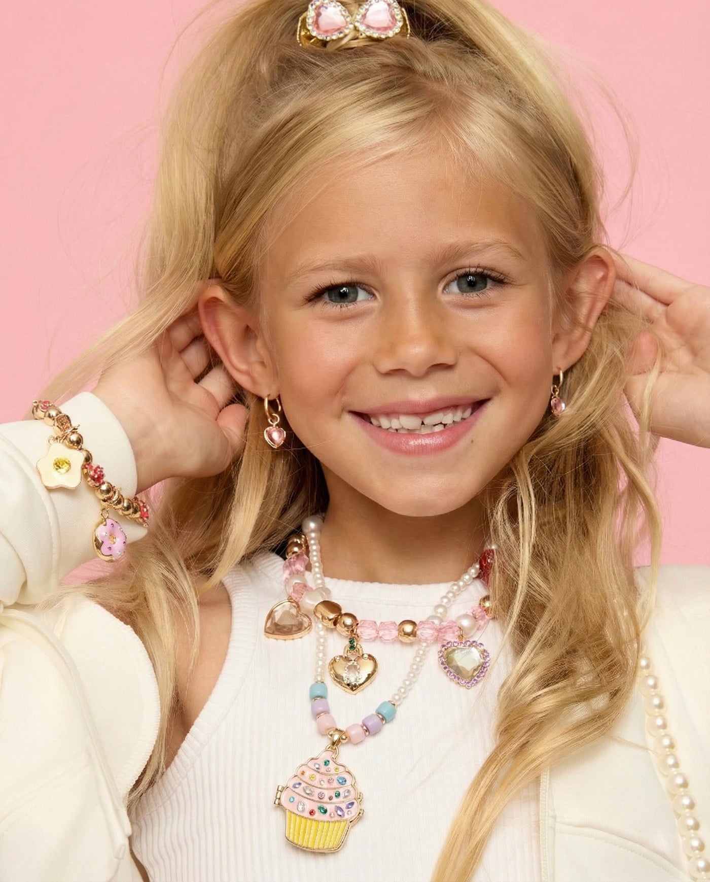 A young girl with long blonde hair smiles in front of a pink background, wearing Super Smalls pastel jewelry and the Cupcake Lipgloss Locket pendant.