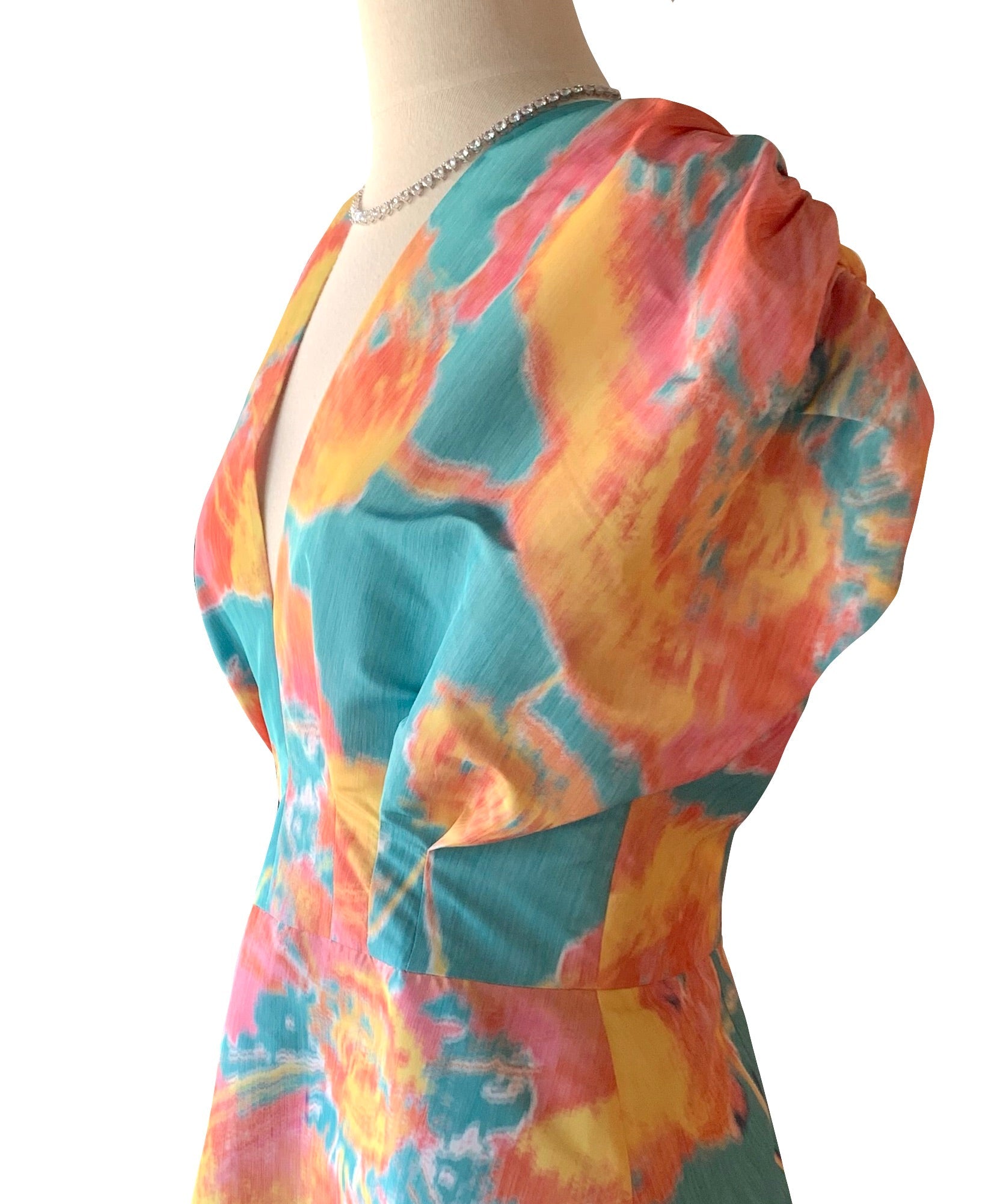 A mannequin dressed in the vibrant Lela Rose Isabel Dress with a deep V-neckline and cap sleeves. The silky v-neck dress features a painterly print with a mix of blue, pink, and yellow tie-dye patterns.