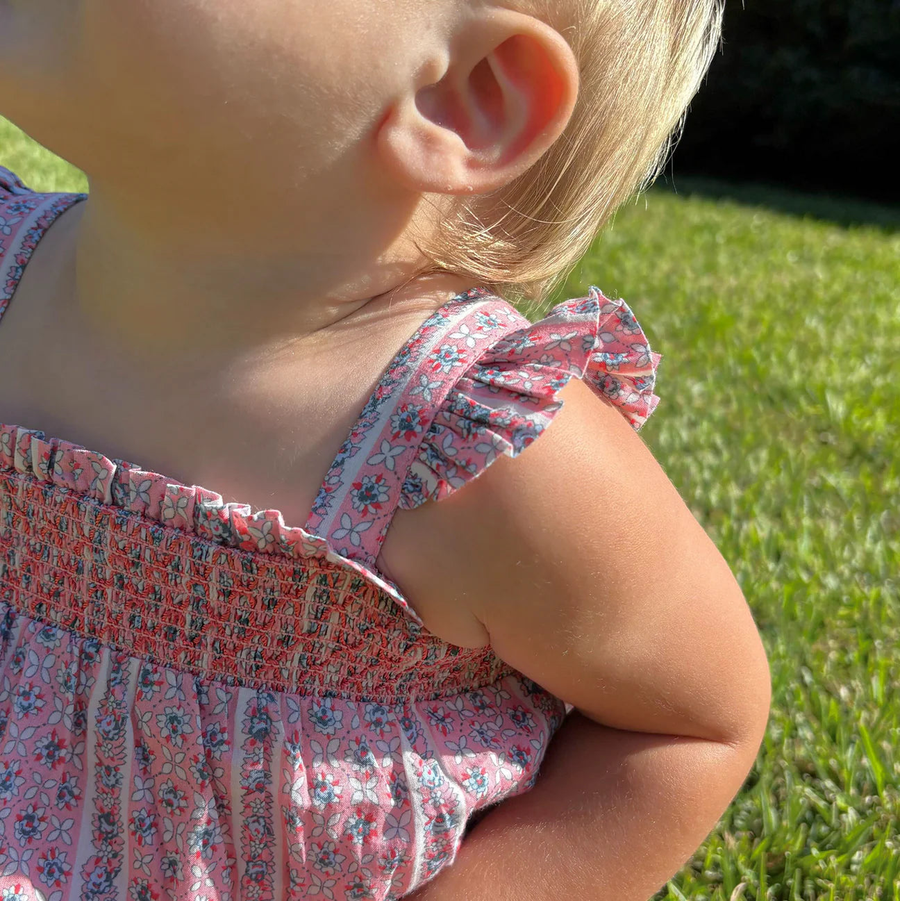 A close-up of a small child wearing the Minnow Baby Girls' Floral Tile Smocked Bubble Romper, standing outdoors on green grass in bright sunlight.