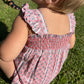 A young child wears the Minnow Baby Girls' Floral Tile Smocked Bubble Romper while sitting on green grass with sunlight casting shadows.