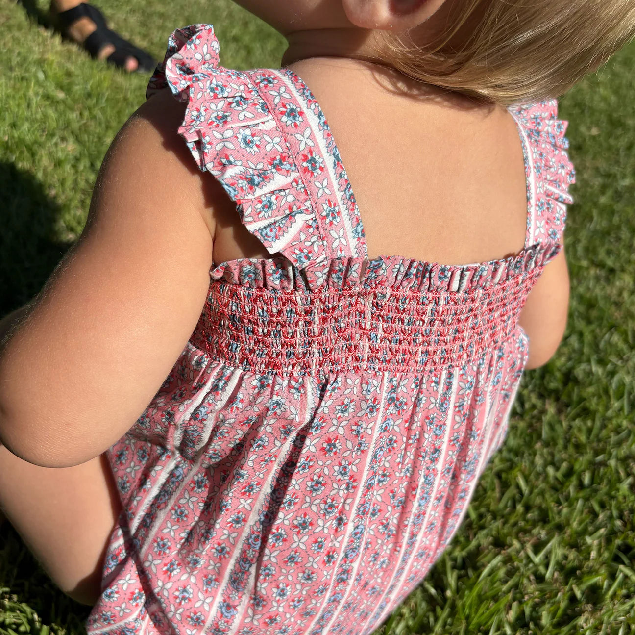A young child wears the Minnow Baby Girls' Floral Tile Smocked Bubble Romper while sitting on green grass with sunlight casting shadows.