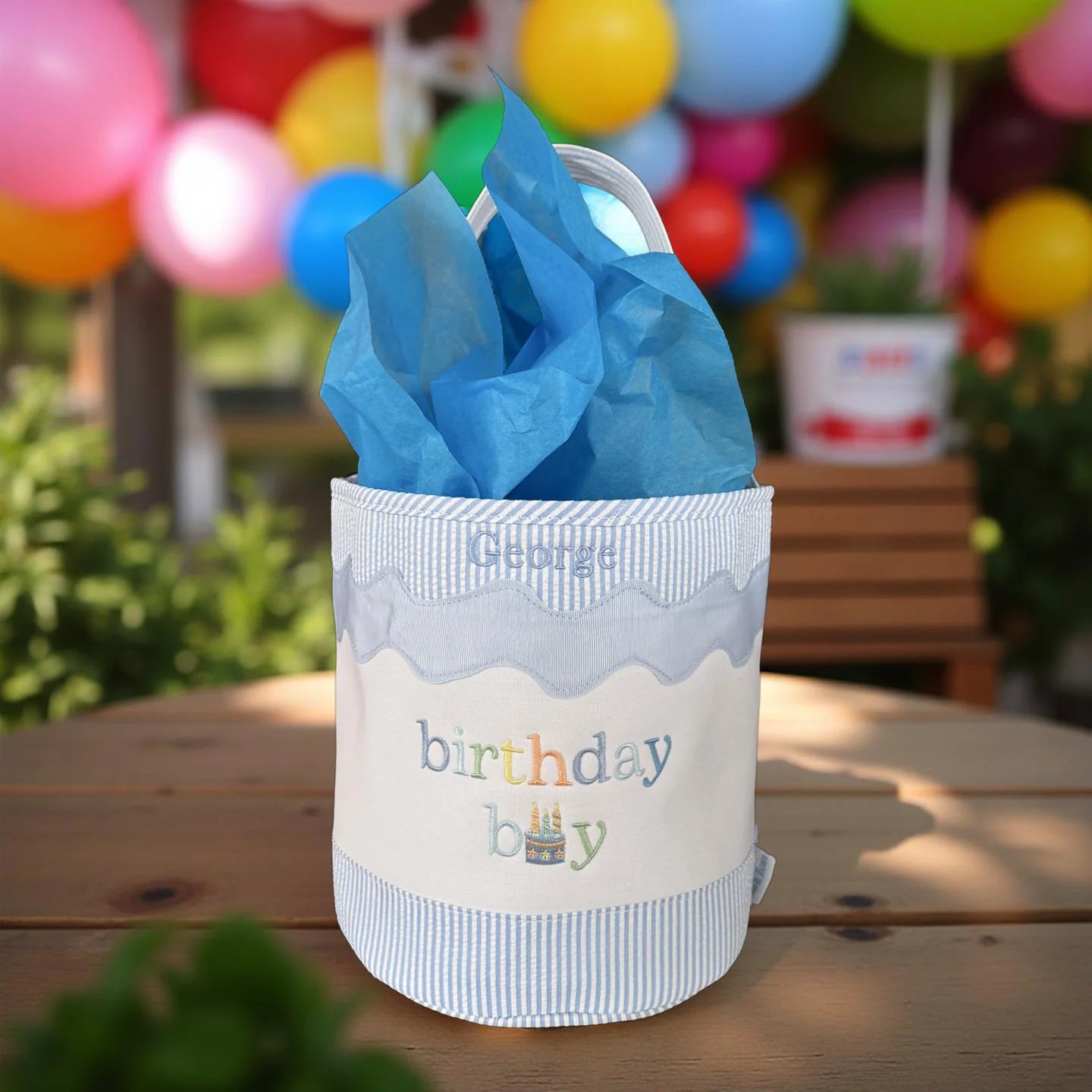 The Bits & Bows Birthday Boy Tote, featuring "birthday boy" lettering and filled with blue tissue paper, sits on a table with colorful balloons—ideal as a giftable bucket or for toy storage.