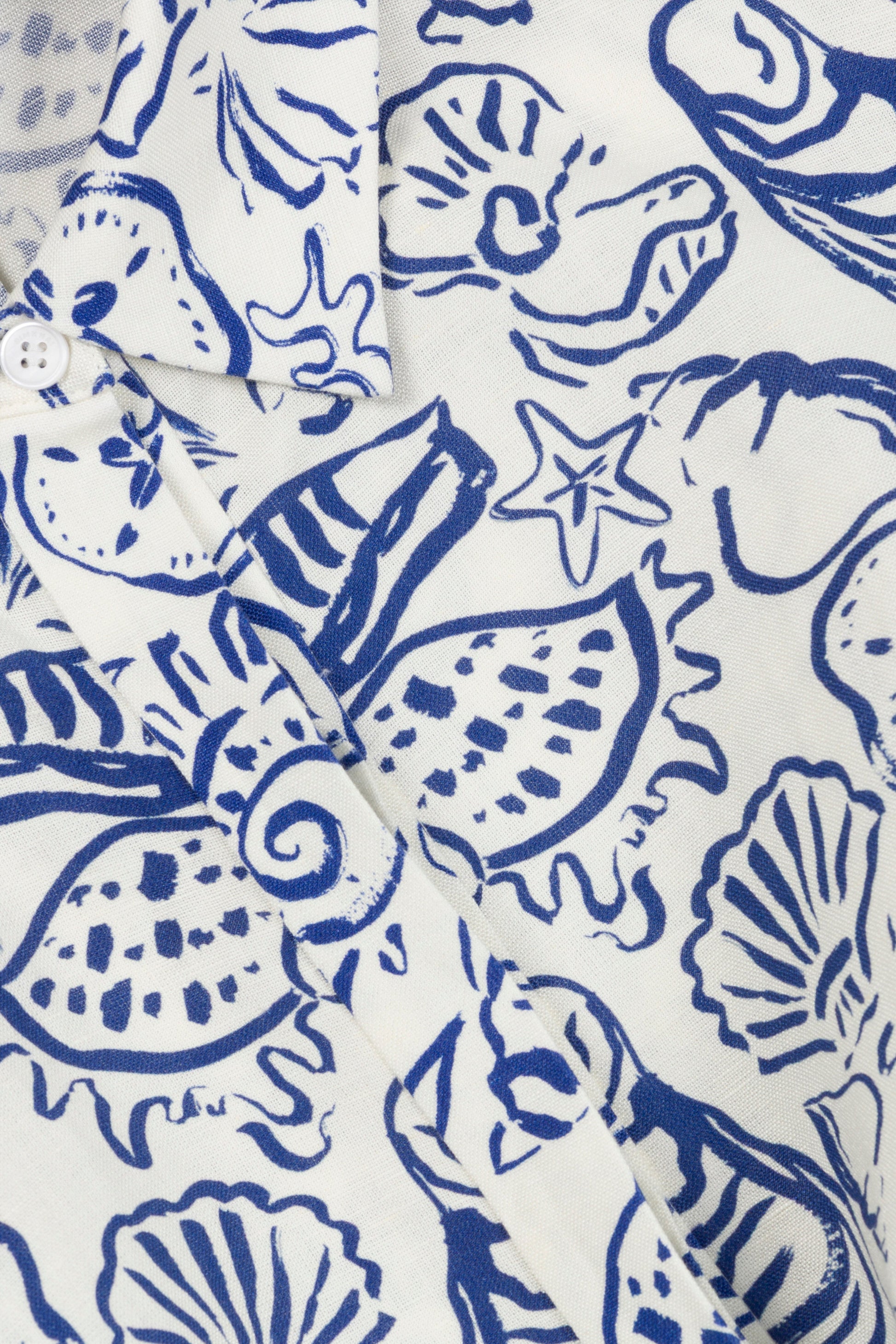 Close-up of the Simkhai Audria Linen Midi Dress, showcasing its blue seashell and starfish print on white fabric with focus on the collar, button detail, and upper chest area.