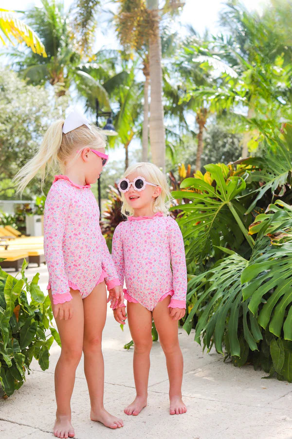 Two young children in matching The Beaufort Bonnet Company Baby Girls and Girls' Sarasota Surf Suits and sunglasses stand barefoot on a path amid tropical plants, highlighting adorable style and sun protection.