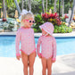 Two young girls by a pool wear matching Baby Girls and Girls' Sarasota Surf Suits from The Beaufort Bonnet Company—one in sunglasses with a bow, the other in a sunhat—enjoying sun protection among palm trees and tropical plants.