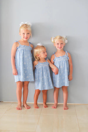Three young girls with blond hair, in matching Girls' Laineys Little Dress by The Beaufort Bonnet Company and white hair bows, stand barefoot against a light gray wall on a tiled floor.