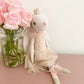 Bijoux the Ballerina Bunny by Mon Ami, wearing a crown and tutu, sits next to pink roses in a glass jar on a light pink surface.