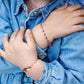 A child in a denim jacket crosses arms, highlighting two beaded bracelets: one red with an 18-carat gold charm and the other multicolored, resembling the Little Gigi Classic Bracelet by Gigi Clozeau.