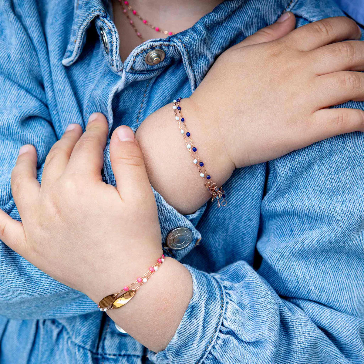 A child in a denim jacket crosses their arms, highlighting two beaded bracelets: a pink and 18-carat gold Gigi Clozeau Little Gigi Classic Bracelet 5.1" and another bracelet in purple and gold with delicate beads.