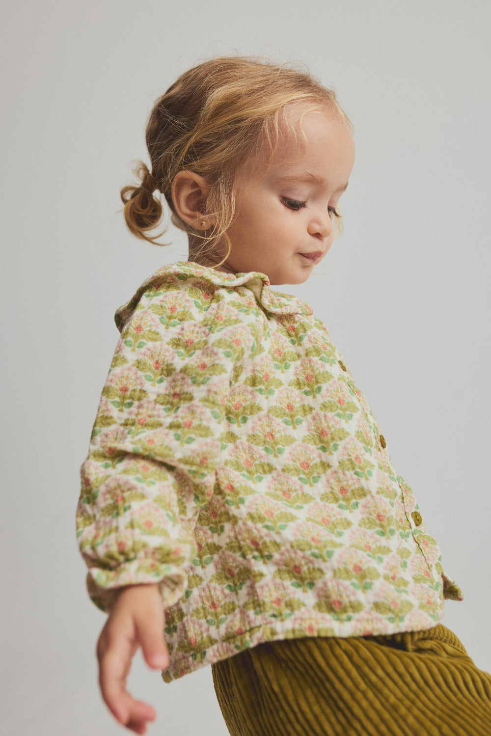 A young toddler girl with light brown hair in pigtails, wearing the Cozmo Baby Girls’ Iris Printed Blouse and mustard corduroy pants, stands against a neutral background.