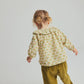 A young child with blonde hair in pigtails faces away, wearing the Cozmo Baby Girls’ Iris Printed Blouse—a patterned long-sleeve floral top—paired with mustard yellow corduroy pants, standing against a plain light background for a stylish everyday toddler look.