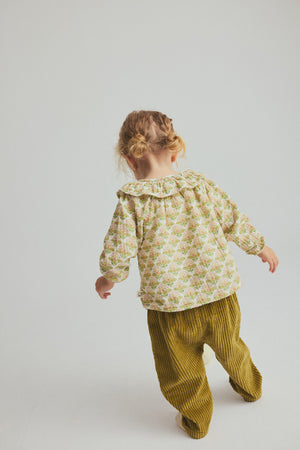 A young child with blonde hair in pigtails faces away, wearing the Cozmo Baby Girls’ Iris Printed Blouse—a patterned long-sleeve floral top—paired with mustard yellow corduroy pants, standing against a plain light background for a stylish everyday toddler look.