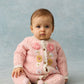 A baby wears the Louise Misha Baby Girls’ Drilia Overalls—pink knit with handmade floral embroidery and scalloped finishes—while sitting on a light blue surface against a matching blue background.
