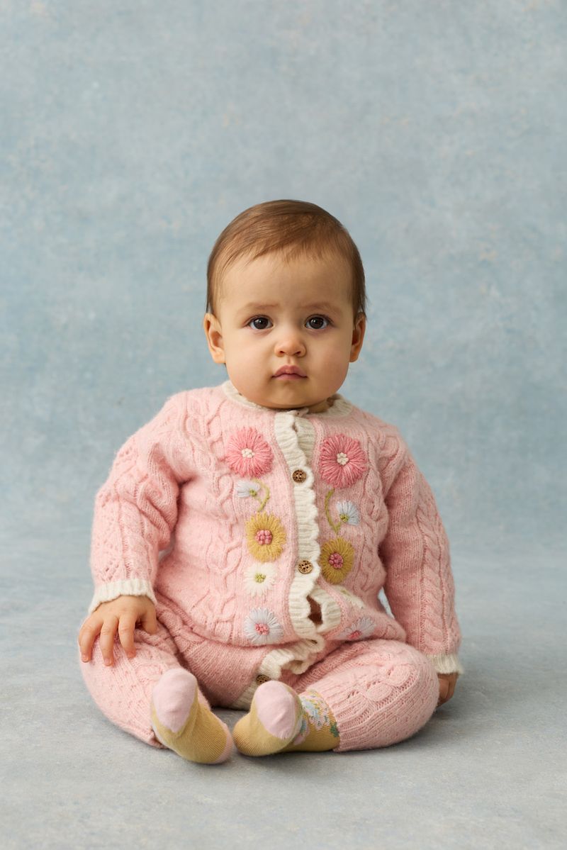 A baby wears the Louise Misha Baby Girls’ Drilia Overalls—pink knit with handmade floral embroidery and scalloped finishes—while sitting on a light blue surface against a matching blue background.