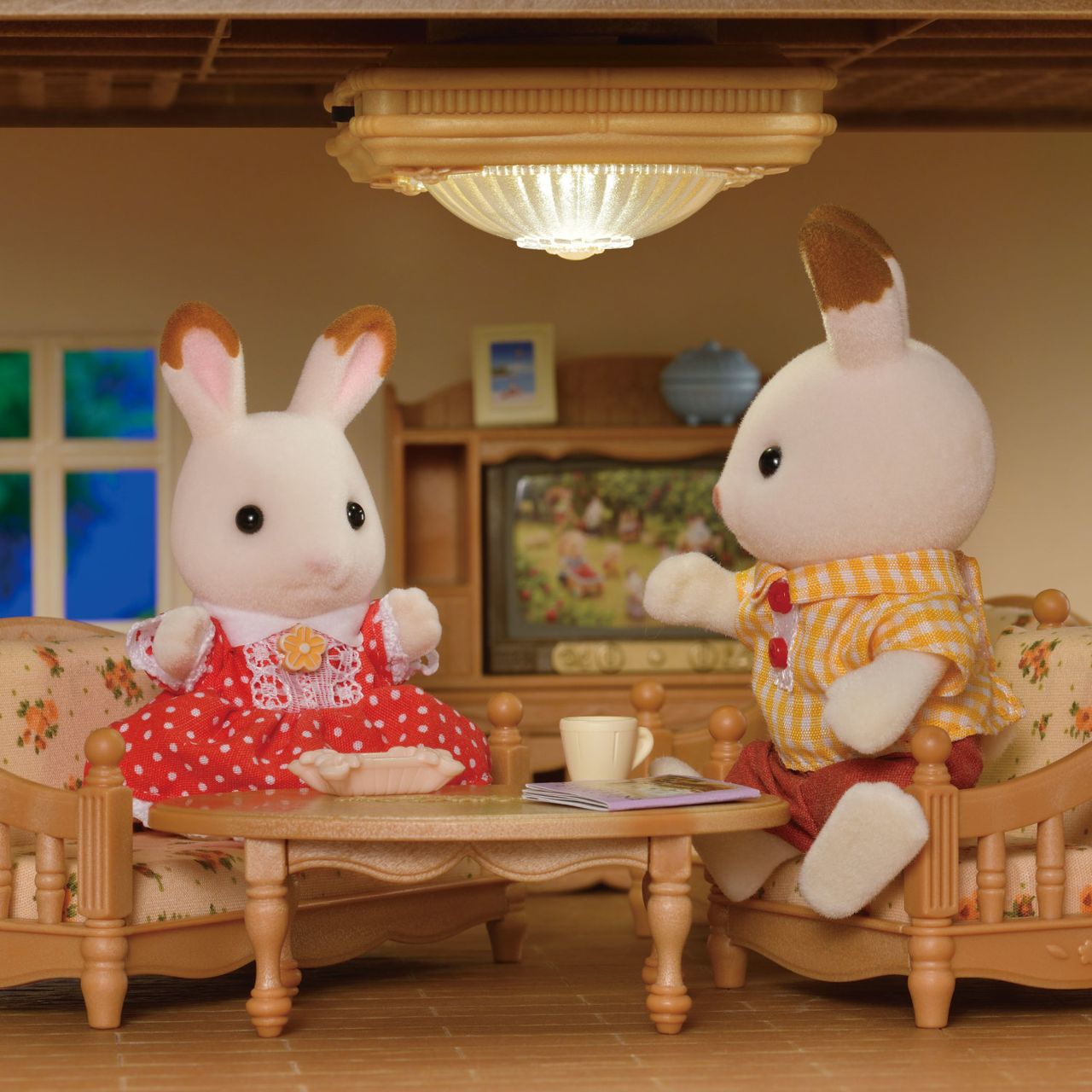 Two toy rabbits sit on floral sofas around a coffee table with a mug and plate, inside a cozy room accented by charming dollhouse décor—a TV, lamp, window, and decorative items complete the scene. The Calico Critters Ceiling Light adds an extra touch of warmth to this inviting miniature setting.