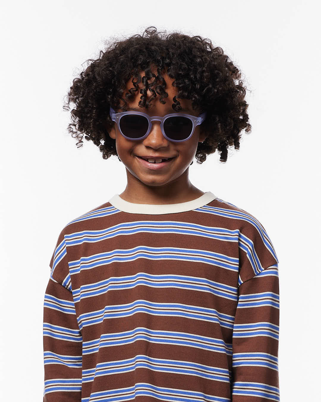 A child with curly hair smiles at the camera while wearing IZIPIZI Kids’ #C Polarized Sunglasses and a long-sleeve shirt featuring brown, blue, and white stripes, standing against a plain white background.