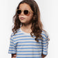 A young child with long brown hair wears IZIPIZI Kids’ #E Polarized Sunglasses, which feature a lightweight tan frame, paired with a white t-shirt with blue stripes, standing against a plain white background.
