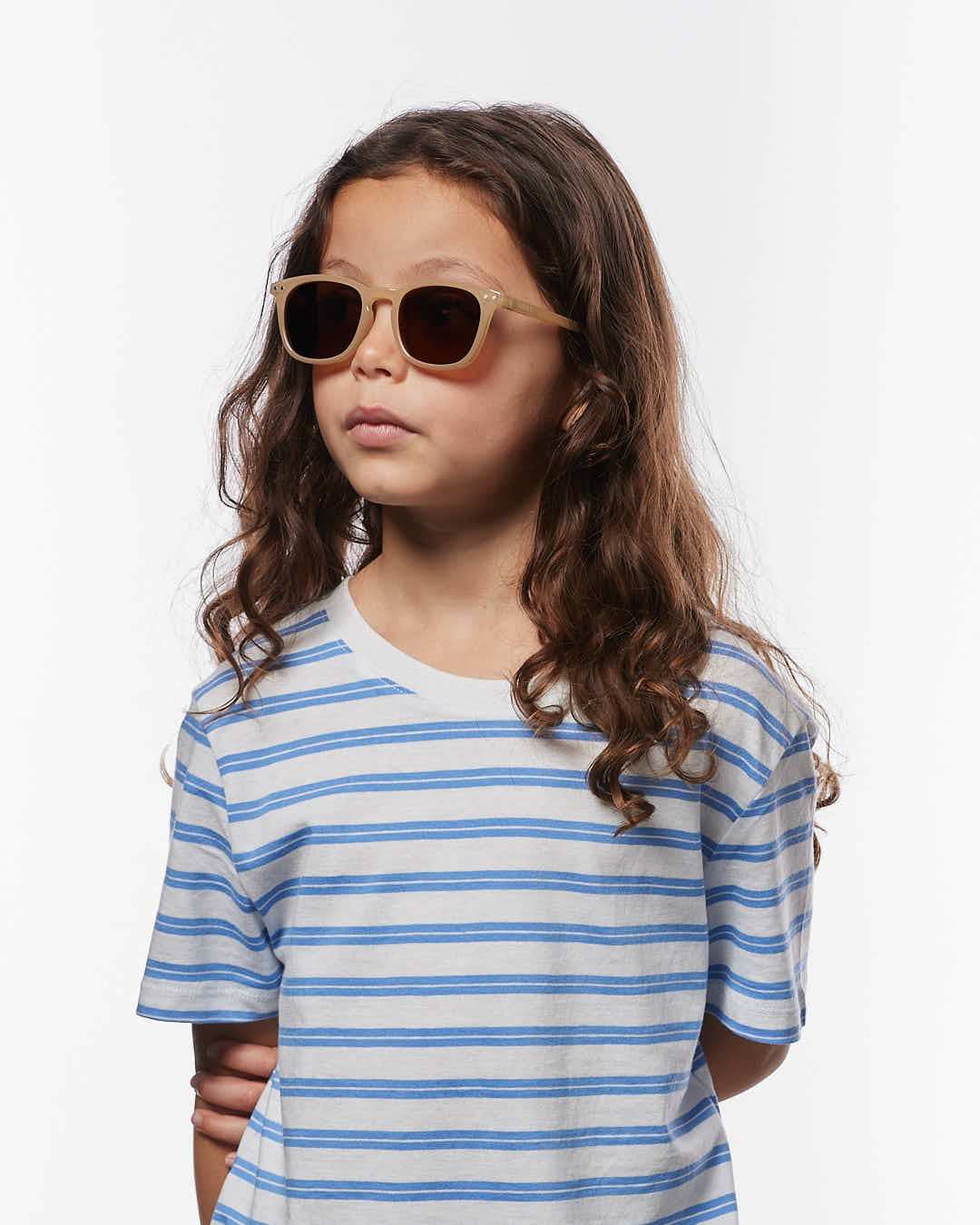A young child with long brown hair wears IZIPIZI Kids’ #E Polarized Sunglasses, which feature a lightweight tan frame, paired with a white t-shirt with blue stripes, standing against a plain white background.