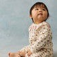 A baby sits on the floor looking upward, wearing Louise Misha’s Girls’ Ysali Sweatpants—soft fleece jogging pants with an elasticated waist and a charming cream-colored floral pattern—set against a light blue background.