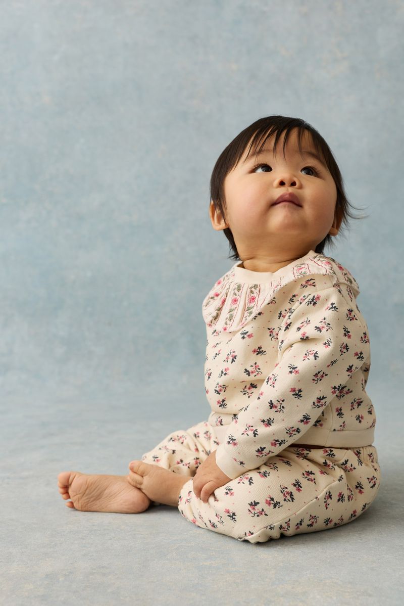 A baby sits on the floor looking upward, wearing Louise Misha’s Girls’ Ysali Sweatpants—soft fleece jogging pants with an elasticated waist and a charming cream-colored floral pattern—set against a light blue background.
