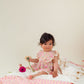 A young child in a Louise Misha Baby Girls' Lena Romper sits on a striped blanket, gently holding two flowers.