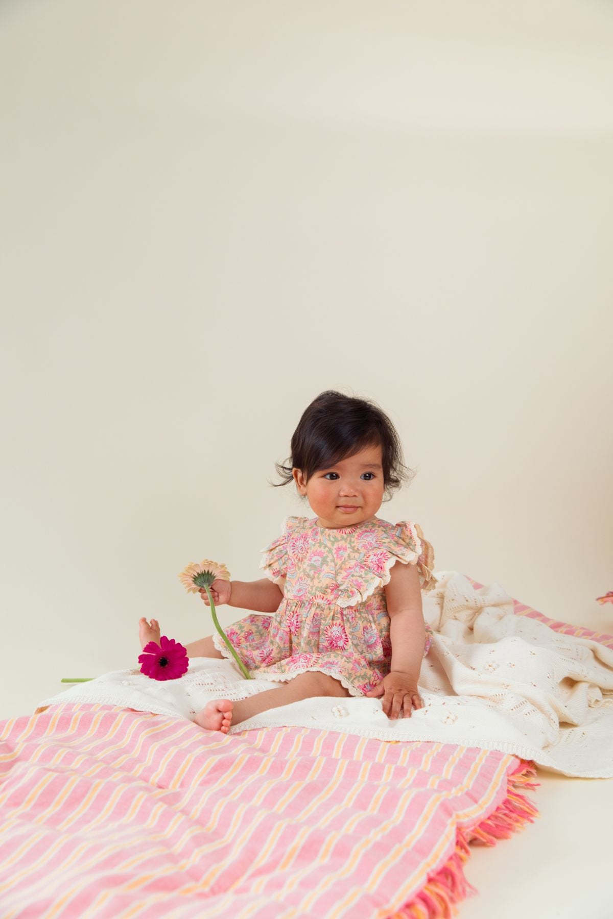 A young child in a Louise Misha Baby Girls' Lena Romper sits on a striped blanket, gently holding two flowers.