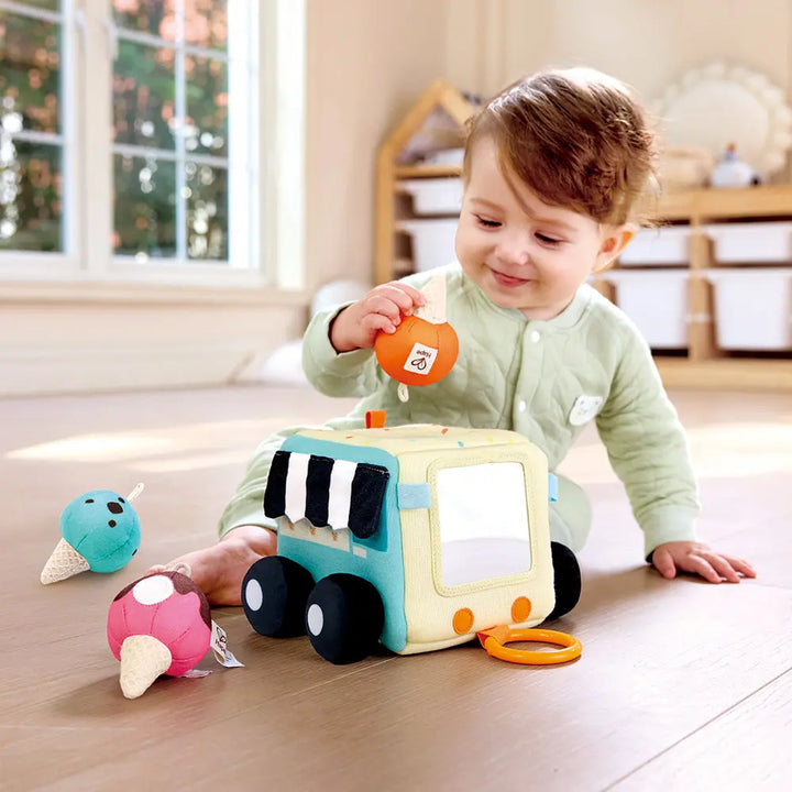 A toddler in green pajamas embarks on a sensory adventure, playing with the Hape Ice Cream Activity Cube on a light-colored floor in a sunlit room.