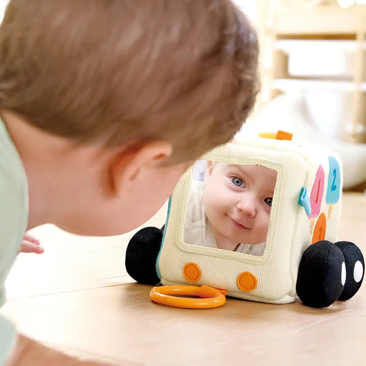 A baby gazes at their reflection in a mirror attached to the Hape Ice Cream Activity Cube, delighting in a sensory adventure as they explore its vibrant colors and engaging textures on the light wood floor.