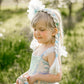 A young child in a blue sparkly dress stands outdoors in the sunlight, wearing the Great Pretenders Ice Queen Princess Hair Braid headband and holding a small toy, with blurred trees in the background.