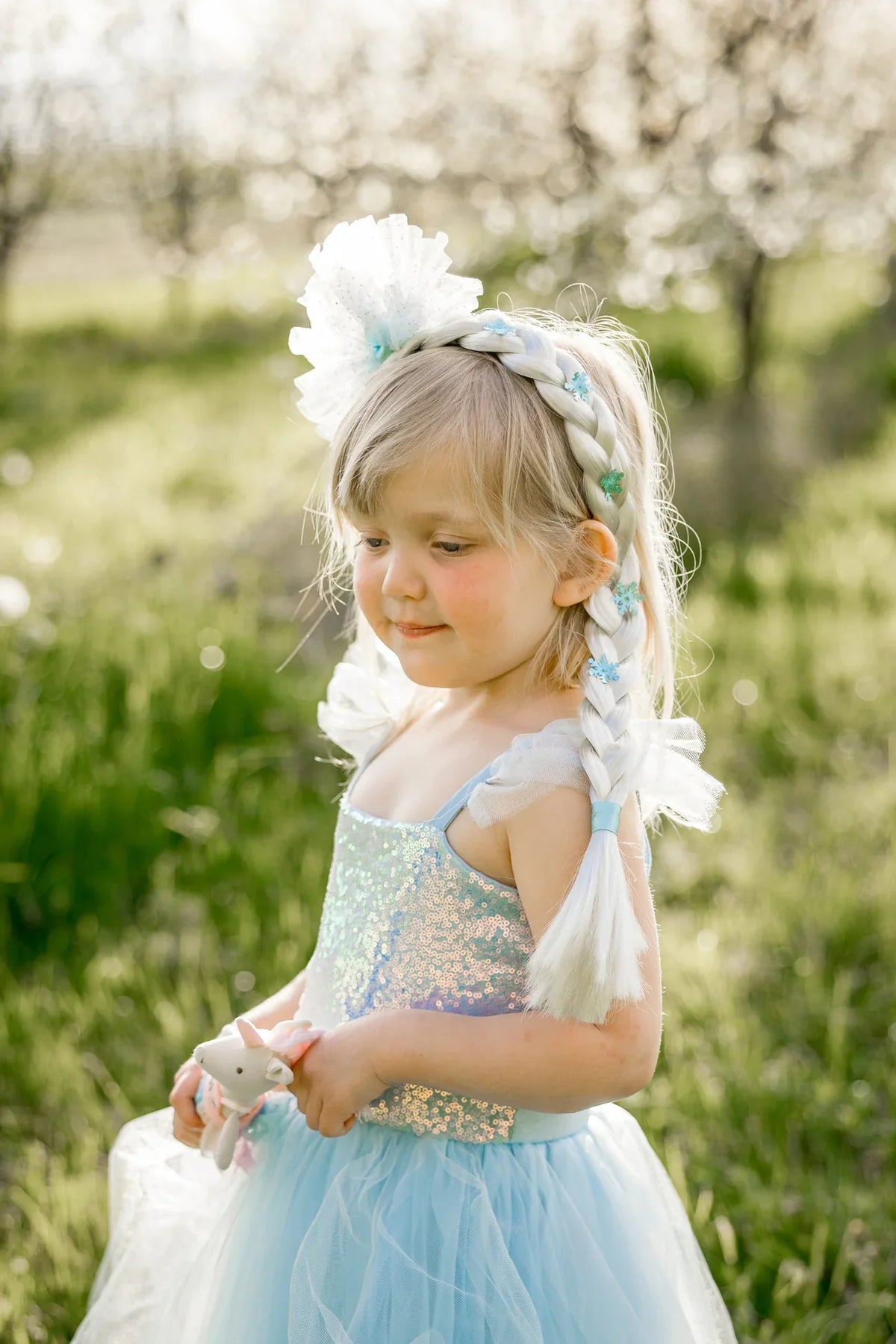 A young child in a blue sparkly dress stands outdoors in the sunlight, wearing the Great Pretenders Ice Queen Princess Hair Braid headband and holding a small toy, with blurred trees in the background.