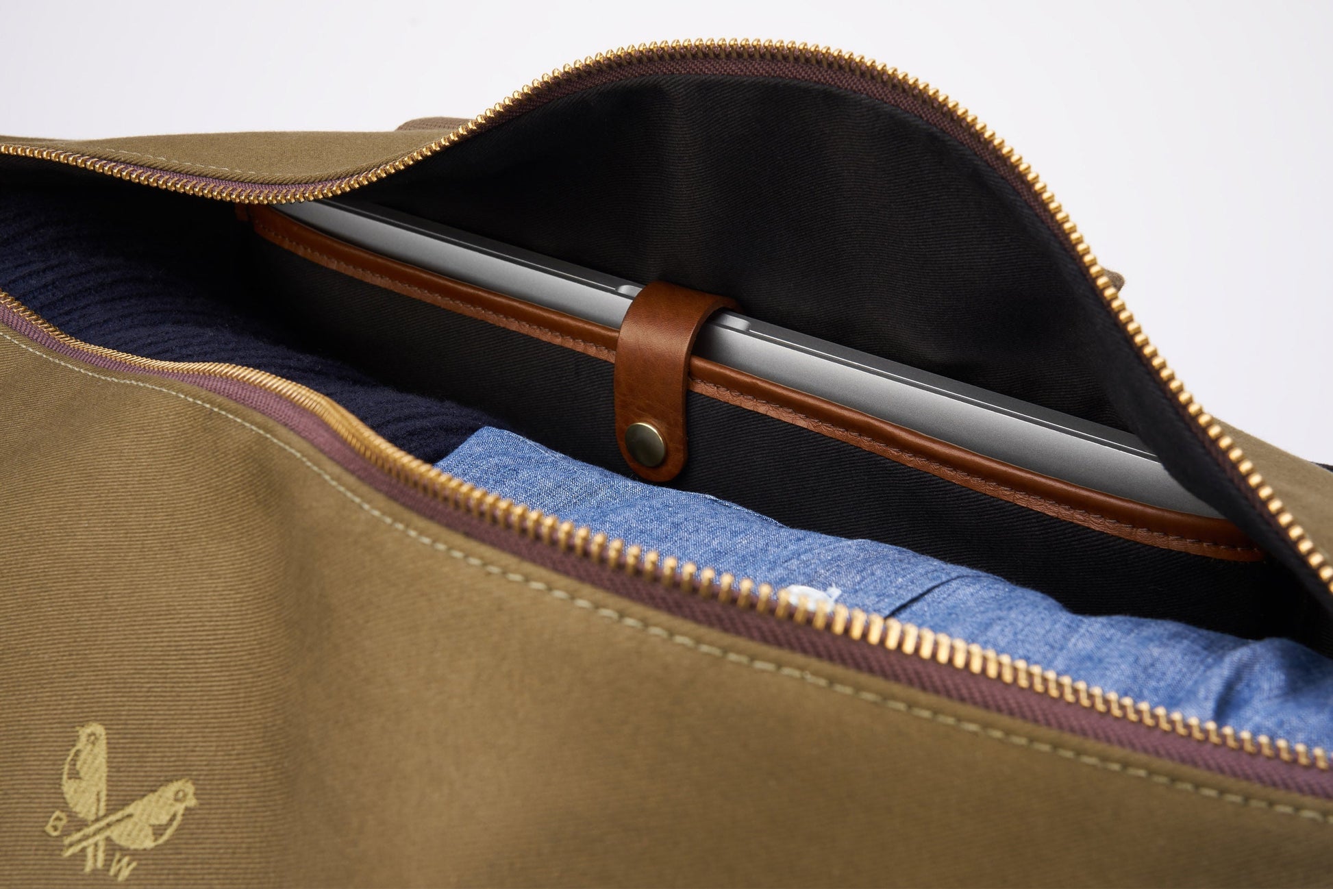 A laptop secured inside a partially open brown Bennett Winch Canvas Weekender bag, crafted from waterproof canvas, with folded blue clothing visible beneath it.