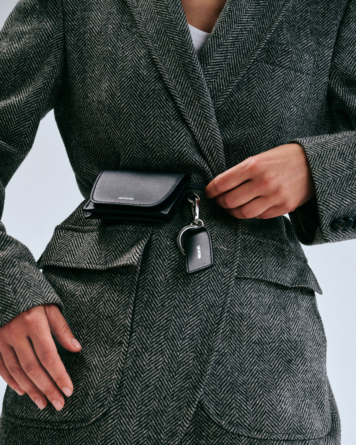 A person in a herringbone blazer attaches the multipurpose Lie Studio Stella Wallet and branded keychain—crafted from Italian cow leather—to their jacket belt area.