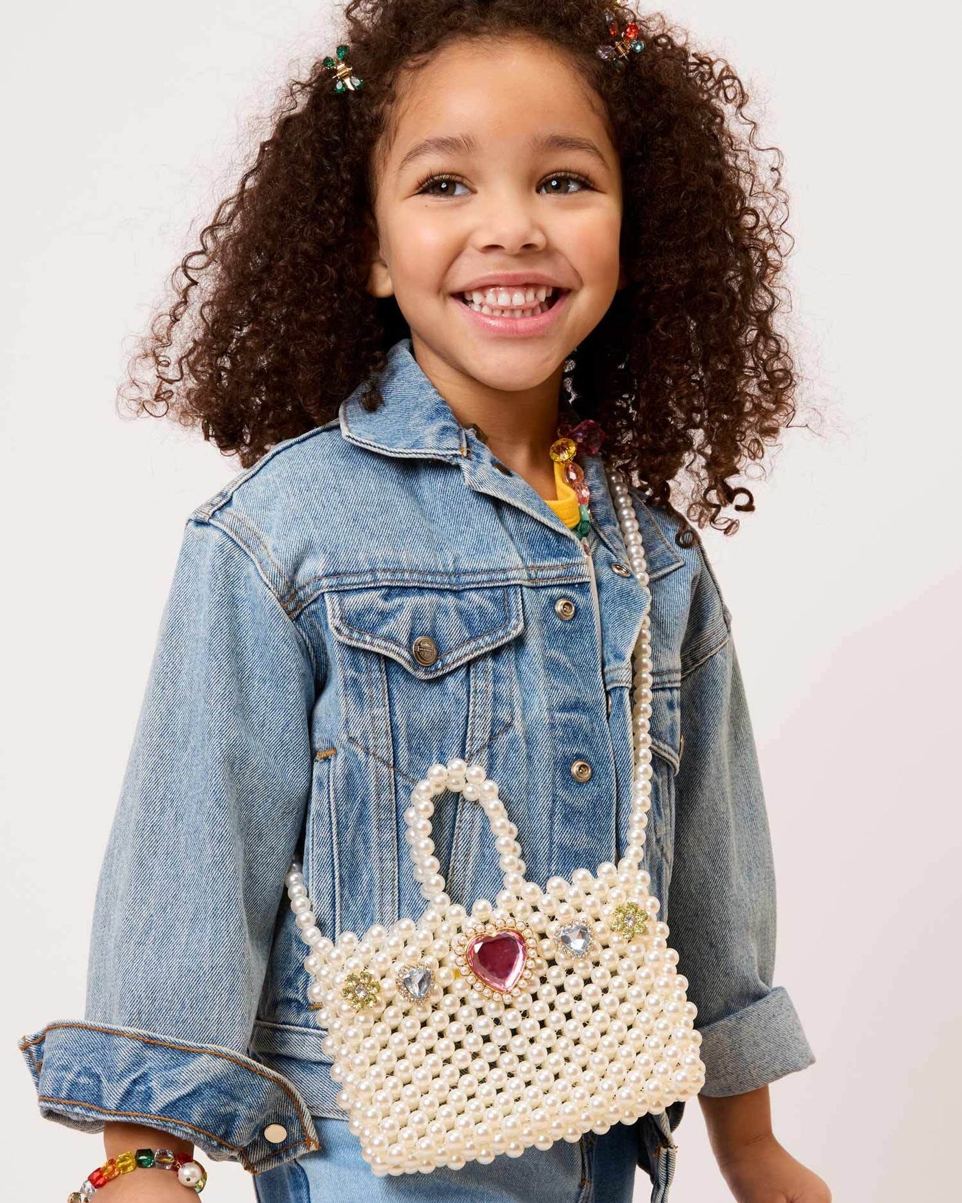 A young girl smiles in a denim jacket, carrying the Super Smalls Pearl of the Party Handbag—a chic kids’ accessory with pearls and a heart-shaped jewel—posed against a plain backdrop.
