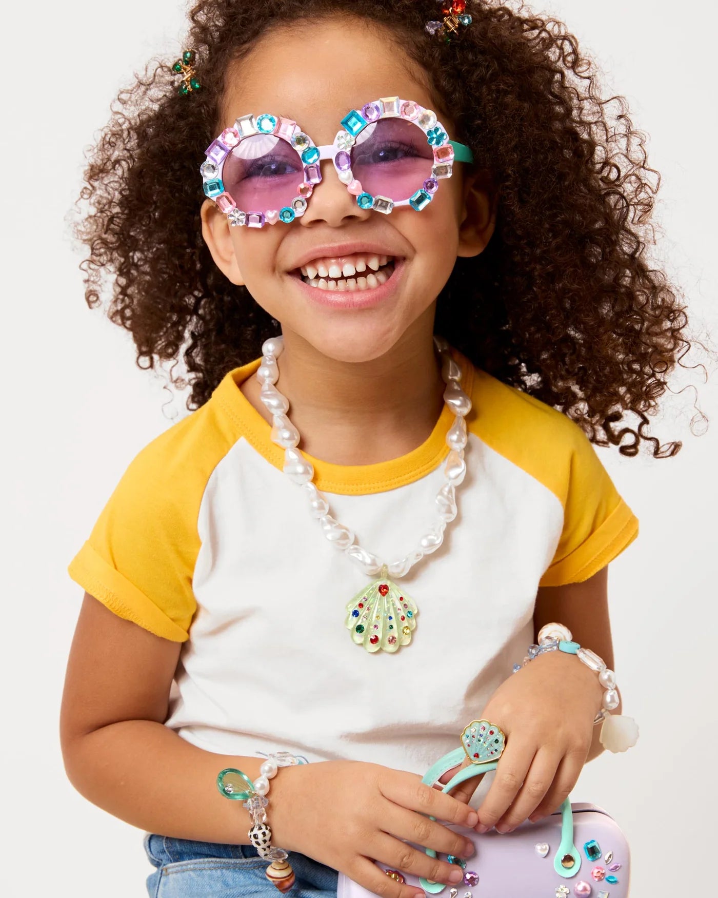 Smiling child with curly hair wears Super Smalls Mermaid Splash & Sparkle Gemify Sunglasses & Case, yellow and white top, chunky necklace, bracelets, and holds a small purple purse—great inspiration for kids’ crafting or DIY gem projects.