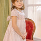A young girl wears the Antoinette Baby Girls' and Girls' Isadora Short Sleeve Floral Smocked Dress, standing indoors beside a red upholstered chair with a window and green curtains in the background.