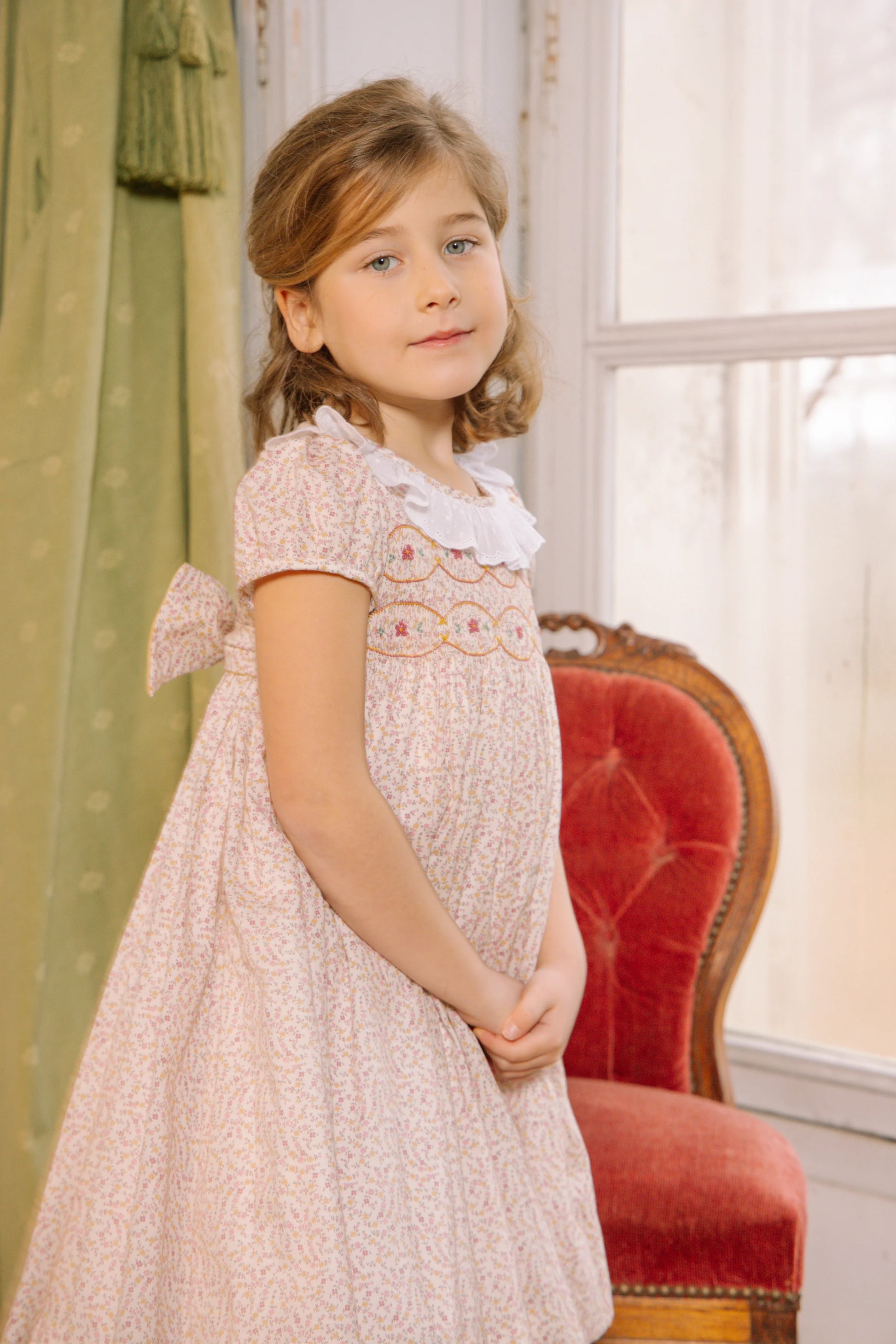 A young girl wears the Antoinette Baby Girls' and Girls' Isadora Short Sleeve Floral Smocked Dress, standing indoors beside a red upholstered chair with a window and green curtains in the background.