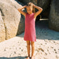 A young girl in the Feather 4 Arrow Girls’ Island Escape Dress stands barefoot on sandy ground near large rocks, her hands resting on her head.