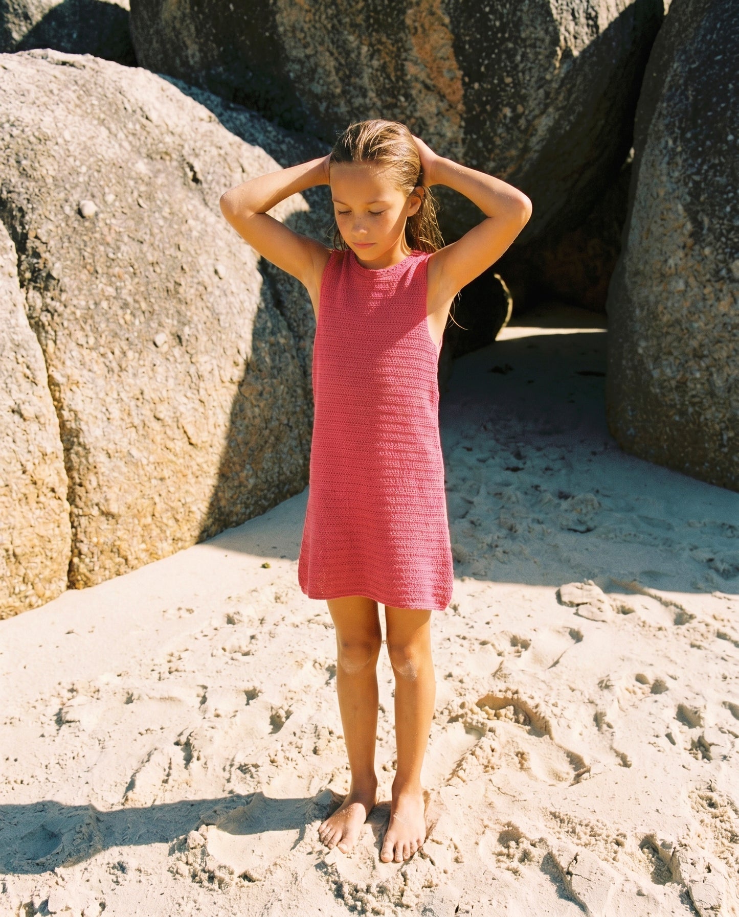 A young girl in the Feather 4 Arrow Girls’ Island Escape Dress stands barefoot on sandy ground near large rocks, her hands resting on her head.