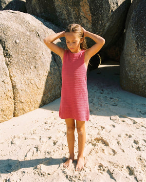 A young girl in the Feather 4 Arrow Girls’ Island Escape Dress stands barefoot on sandy ground near large rocks, her hands resting on her head.