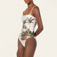 Woman wearing the Durazno Arena One Piece by Agua By Agua Bendita, a UPF 50 swimsuit with thin straps and a tropical palm tree island print, crafted from recycled polyester, standing against a plain background.