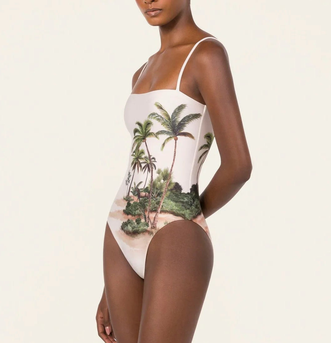 Woman wearing the Durazno Arena One Piece by Agua By Agua Bendita, a UPF 50 swimsuit with thin straps and a tropical palm tree island print, crafted from recycled polyester, standing against a plain background.