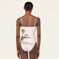 A woman faces away, wearing the Durazno Arena One Piece by Agua By Agua Bendita—a white swimsuit with thin straps, made from recycled polyester and detailed with a beach scene on the lower back.