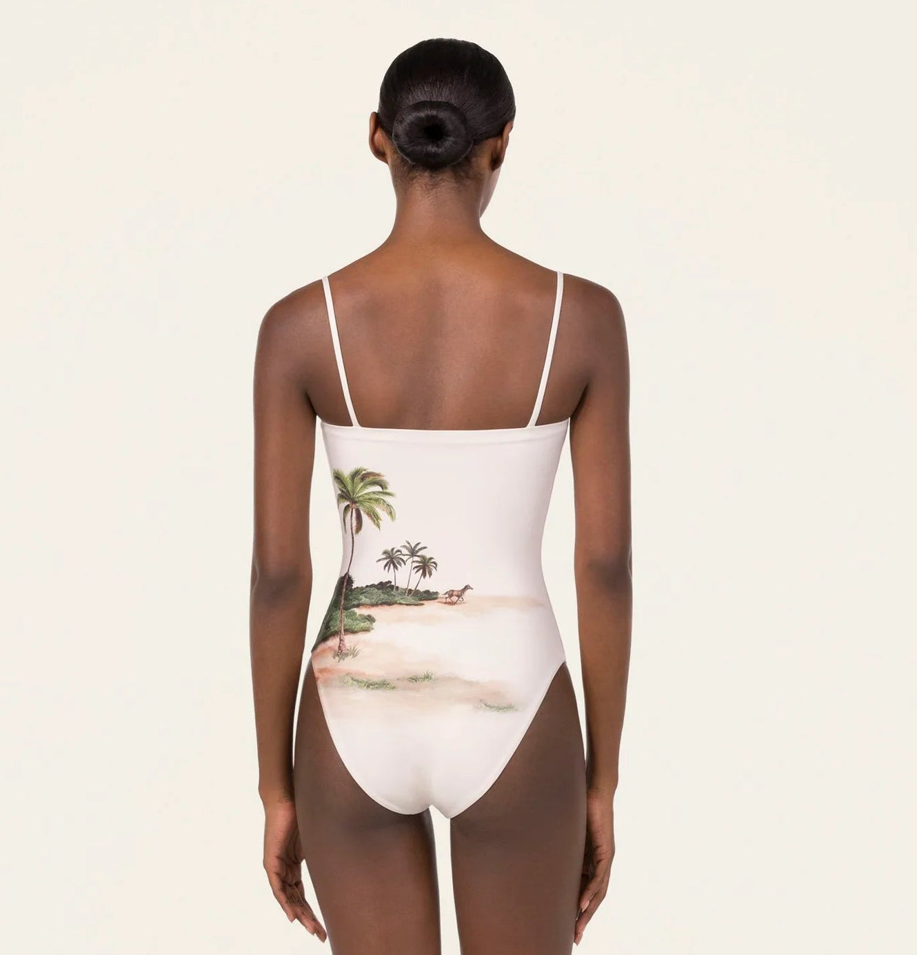 A woman faces away, wearing the Durazno Arena One Piece by Agua By Agua Bendita—a white swimsuit with thin straps, made from recycled polyester and detailed with a beach scene on the lower back.