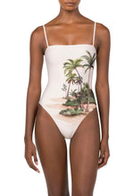 A woman wears the Durazno Arena One Piece by Agua By Agua Bendita, a white swimsuit made from recycled polyester with a tropical beach and palm tree print, standing against a plain background.