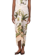 A person wearing the Lavanda Arena Pareo by Agua By Agua Bendita, featuring a tropical palm tree and beach print, paired with a white top and brown flats.