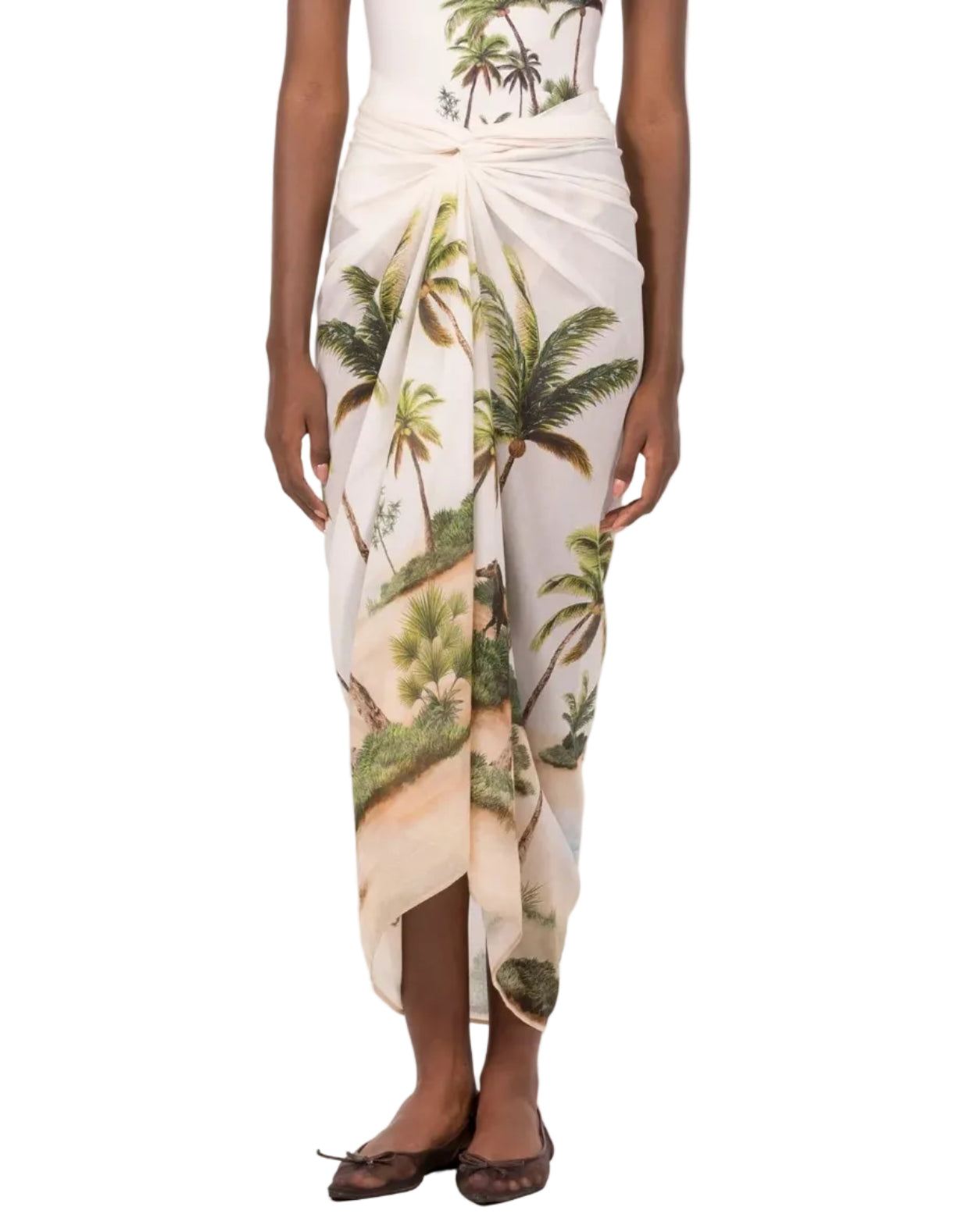 A person wearing the Lavanda Arena Pareo by Agua By Agua Bendita, featuring a tropical palm tree and beach print, paired with a white top and brown flats.