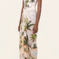 A woman wears the Lavanda Arena Pareo by Agua By Agua Bendita—a white spaghetti-strap dress with a tropical palm tree print, styled as a chic swimsuit cover-up and paired with flat brown sandals.