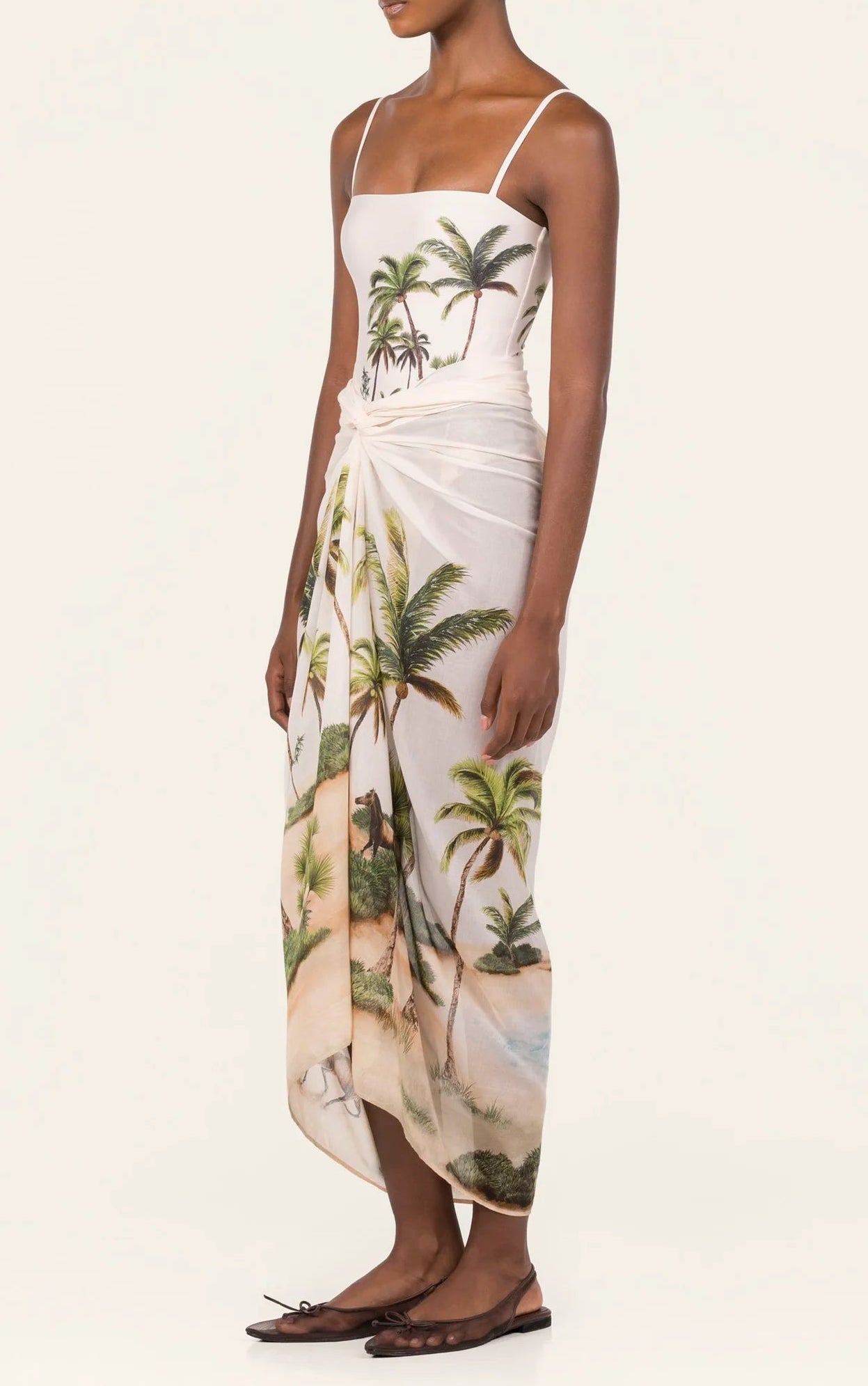 A woman wears the Lavanda Arena Pareo by Agua By Agua Bendita—a white spaghetti-strap dress with a tropical palm tree print, styled as a chic swimsuit cover-up and paired with flat brown sandals.