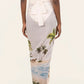 A person stands facing away, wearing the Lavanda Arena Pareo by Agua By Agua Bendita—a white skirt with a beach and palm tree print, tied at the waist and paired with sandals for chic beachwear.