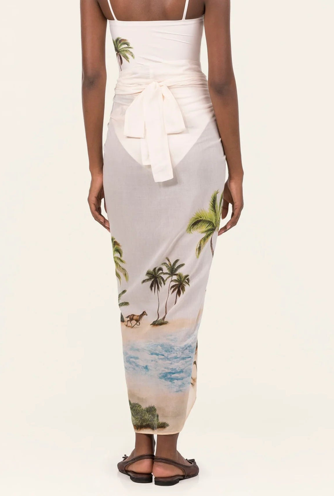 A person stands facing away, wearing the Lavanda Arena Pareo by Agua By Agua Bendita—a white skirt with a beach and palm tree print, tied at the waist and paired with sandals for chic beachwear.